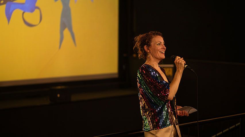 Stacy smiling and speaking into a microphone in front of a cinema screen at the Exit 6 Film Festival