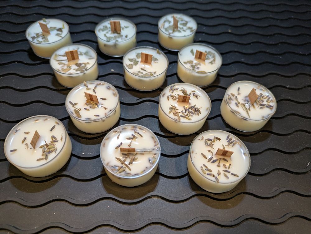 A group of handmade tealight candles with wooden wicks arranged on a black, wavy-textured silicone surface. The candles are creamy white in color and topped with dried lavender and eucalyptus. Each candle is set in a small, round, clear container.

