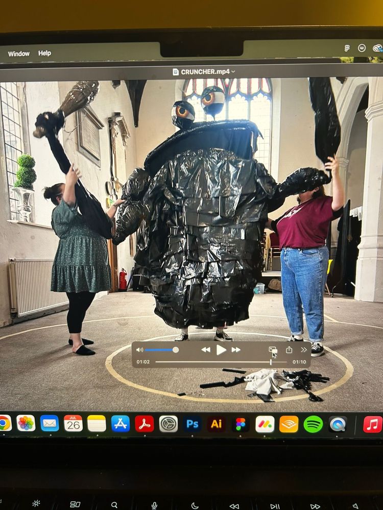A huge trash monster puppet with a person operating each arm, giant yellow balloon sized eyes and a big club. 