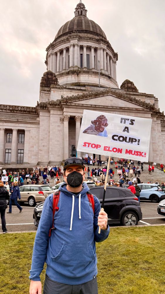 A masked man in front of Olympia, WA capital building. The sign has a picture of Admiral Ackbar and says "It's a coup! Stop Elon Musk"