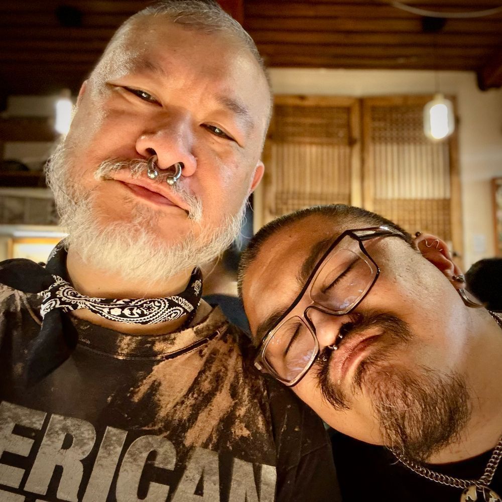 A color photo of two Asian men in warm lighting. The man on the left has a white beard, and is wearing a septum ring, and a black neckerchief with a distressed black shirt. The man on Wright has his head, leaning on the other man’s shoulder, and has black hair, a black goatee, a small septum piercing, several earrings, and glasses.