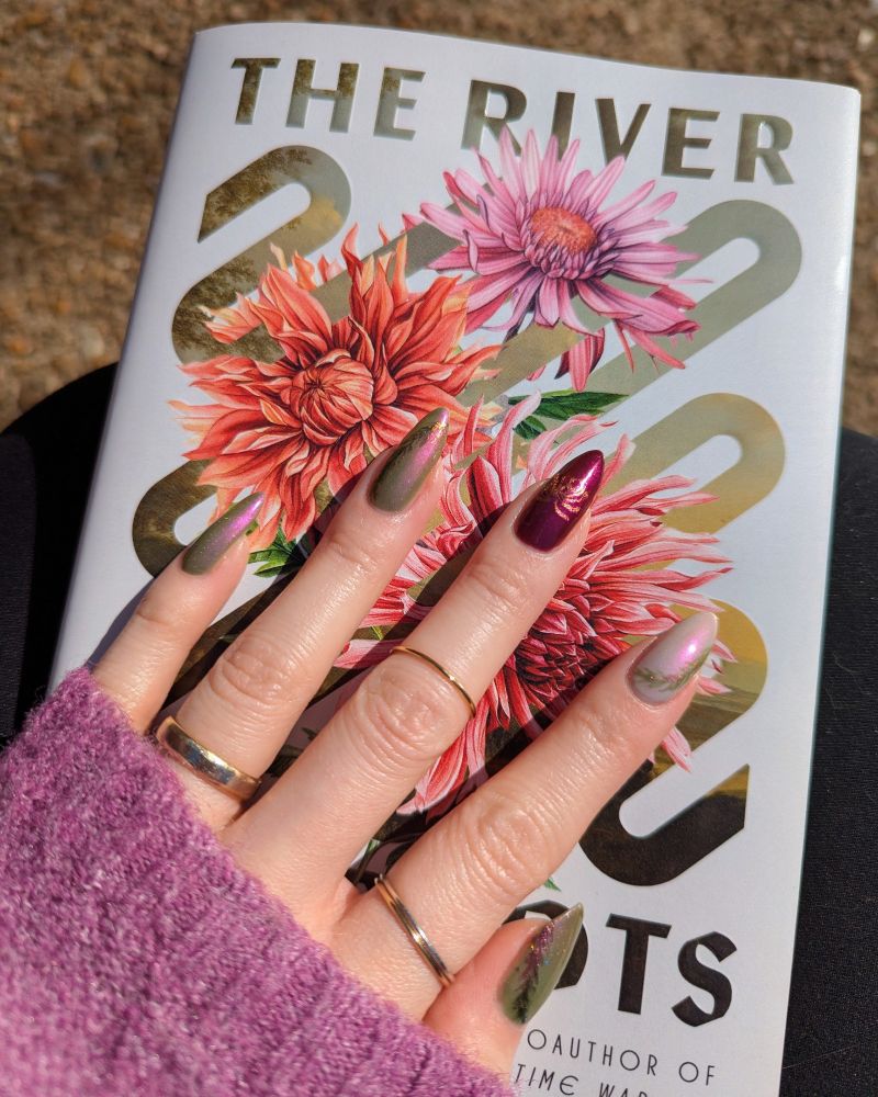 left hand with painted nails placed on top of the cover of the book The River Has Roots by Amal El-Mohtar in direct sunlight