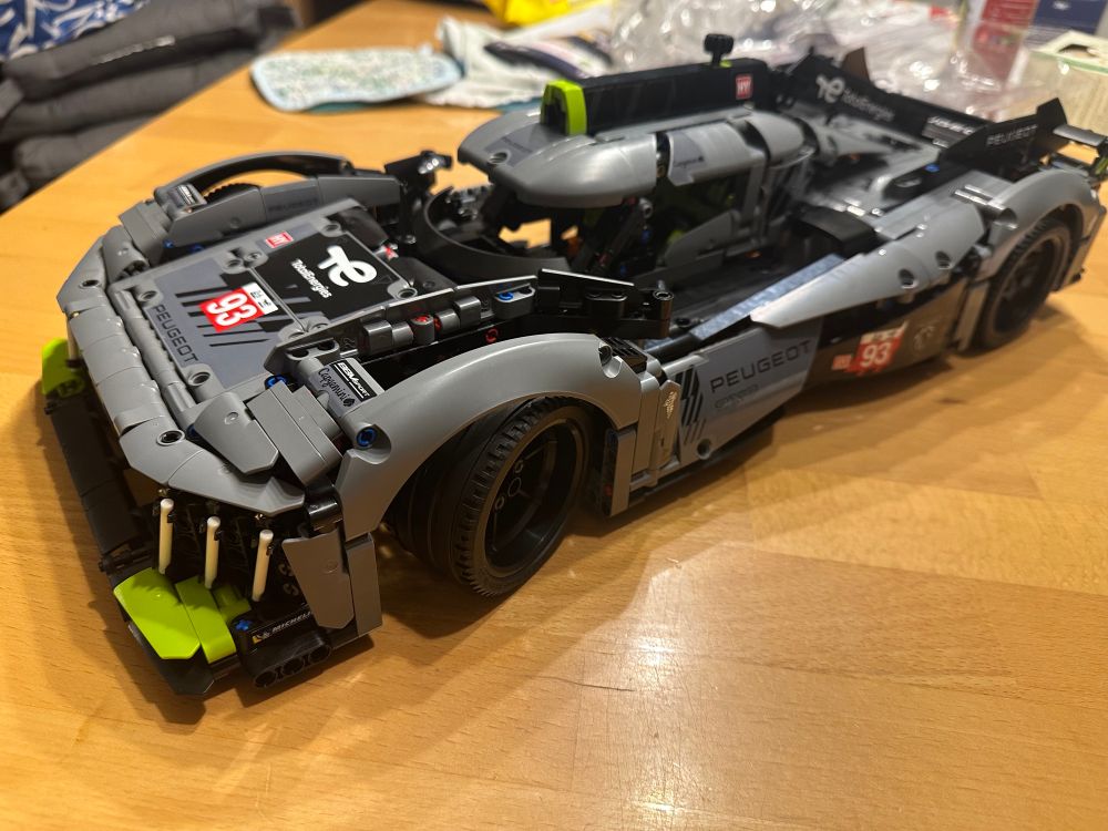 ALT-Text: Ein detailliertes LEGO-Technic-Modell eines Peugeot 9X8 Hypercars in Grau und Schwarz mit grünen Akzenten, das auf einem Holztisch steht. Das Modell zeigt realistische Details wie Aerodynamik-Elemente, Sponsorenlogos, die Startnummer 93 und sichtbare technische Komponenten. Im Hintergrund liegen Verpackungsmaterialien und Haushaltsgegenstände.