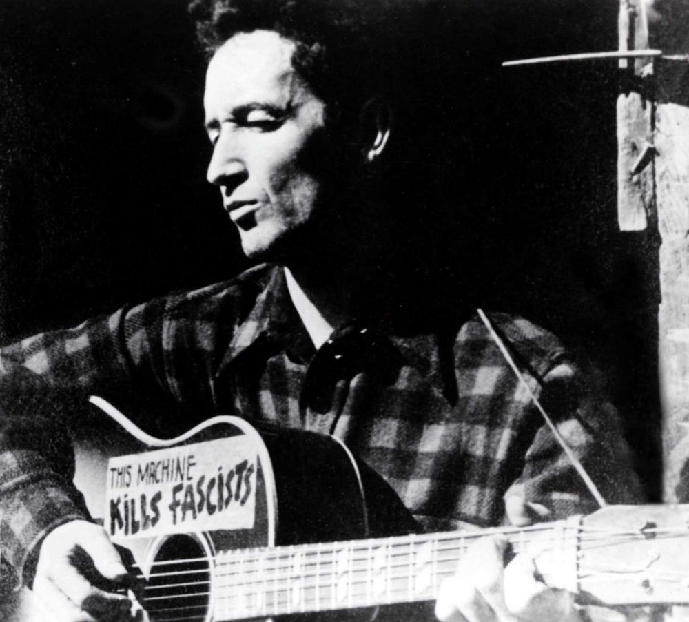 Woody Guthrie with an acoustic guitar that has a sticker that says "this machine kills fascists"