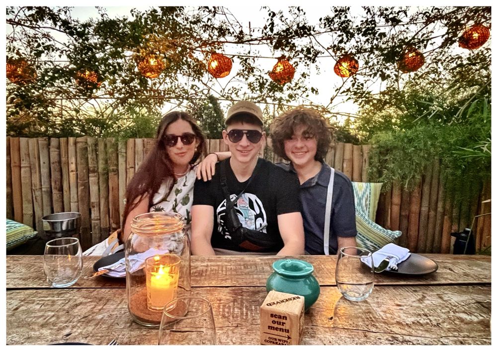 My three kids sitting at a table in Kabana. Fairy lanterns hang in the shrubs behind. 