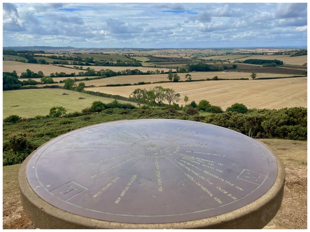 A view from a small hill, looking over the Leicestershire countryside, with fields and hedges making a patchwork scene below. The photo is taken from the edge of a toposcope, which forms the foreground of the photo, with lines radiating out from the centre of its circular top, demonstrating distances to landmarks