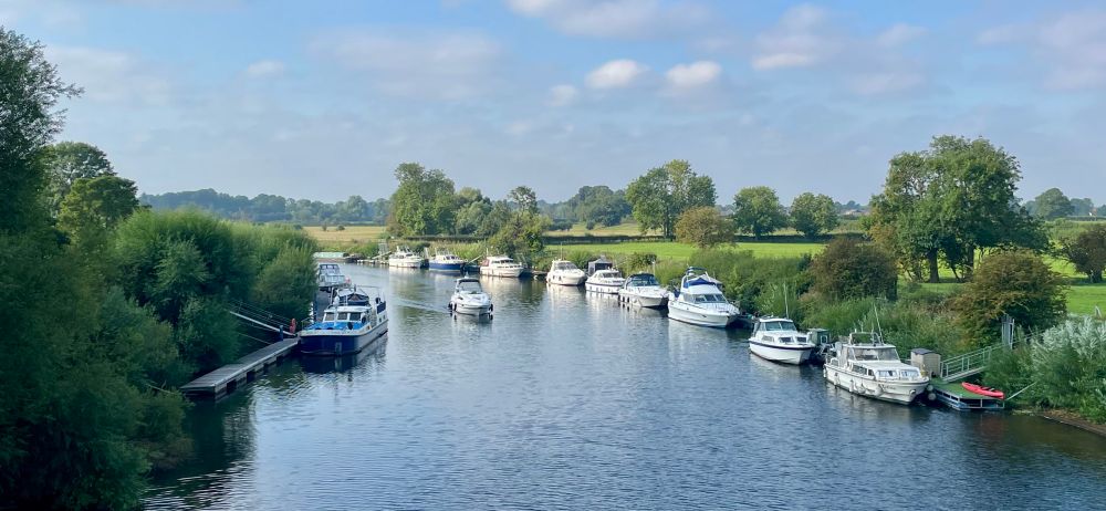 The river Ouse on a sunny day. Boats sides are lined with moored boats and there is one boat travelling towards the camera in the middle of the water. The river is bordered by green fields and trees