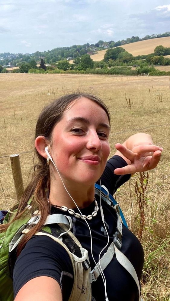 My daughter, Maddy, out on a training walk. She is wearing headphones and a backpack. She has long brown hair that is tied back and she’s making a sideways victory sign and is smiling. 