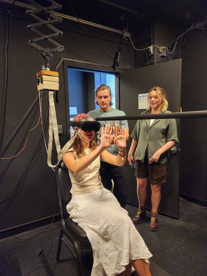A person wearing a virtual reality (VR) headset interacts with their hands in a seated position, demonstrating an immersive VR experience. Two individuals stand nearby, observing and guiding the session in a high-tech setup, featuring equipment and cables in a lab.
