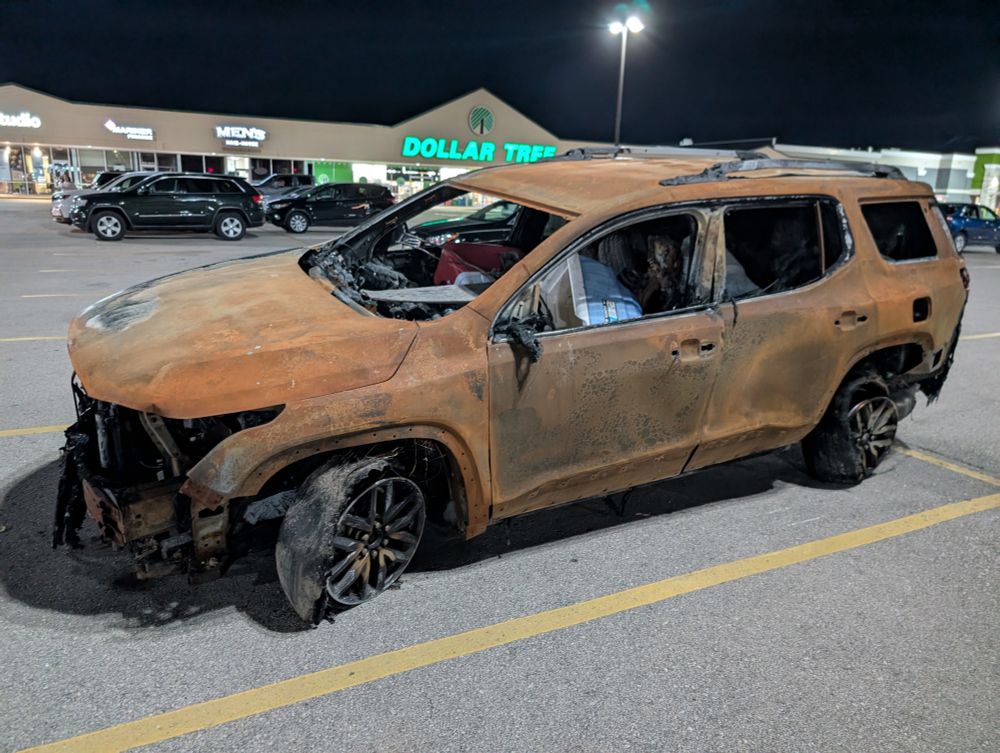 A photo of an SUV in a parking lot. Its tires are melted, windows are busted out, and the whole surface looks crusty and rusted, as if it were burnt out. There are two unburnt coolers in the front seats. Behind it you can see a Dollar Tree.