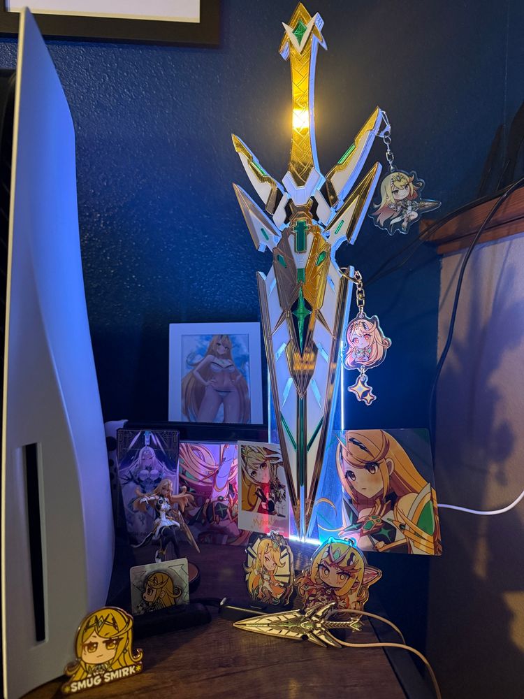The Mythra shrine, with the giant acrylic Aegis lamp and Aegis necklace from Clinkorz, pins, hanging charm, Switch Dock cover, lingerie pin-up, and gacha card from Kamaniki, holo charm from DatCravat, mini-print from Milo Monzon, charm from Alinalal, official amiibo, charm from Kaijucery, and tarot card by tactactak