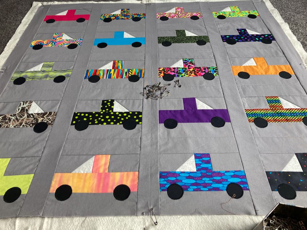 Grey unquilted quilt top on the floor with colourful utes / pickuptrucks made in colourful fabrics. A handful of safety pins are on the centre of the design, as I need to pin baste all the layers together. At the bottom right, is a tin full of safety pins.