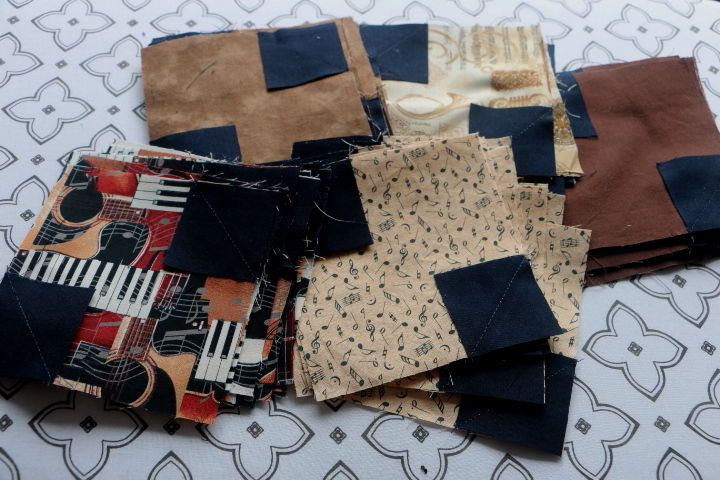 Assorted brown, tan, and music themed fabric squares, with small dark squares sewn to opposite corners. This is called a half snow ball block.