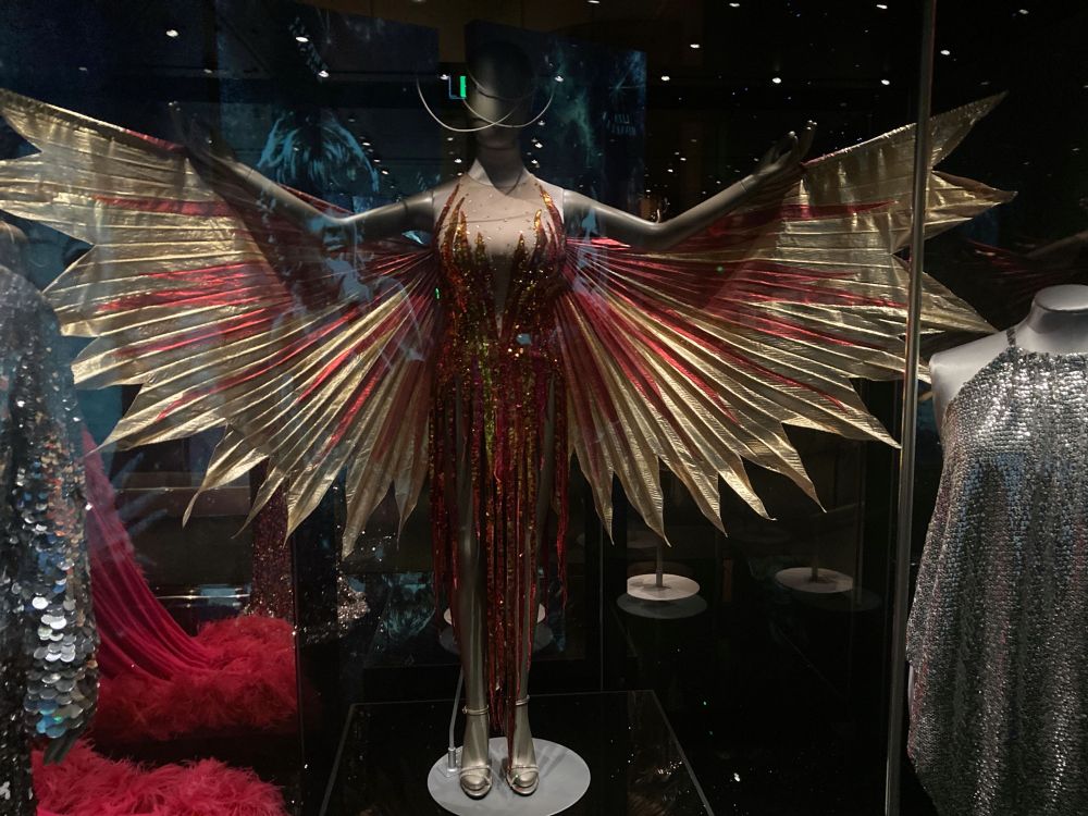 Mannaquin with outstretched arms with gold and red pleated wings . A gold and red dress with fabric creating the impression of flames.