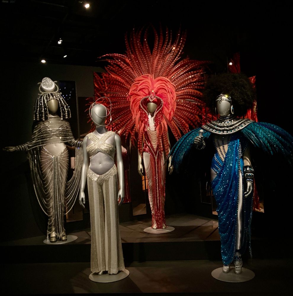 2 mannequinns wearing two gold/silver beaded garments. A red garment is in the background with red feathers extending out like a peacock display. A blue garment is on the right. The colour is heavily beaded, there are feathers protruding like a shawl Lots of shiny beads cover the draped silhoutte.