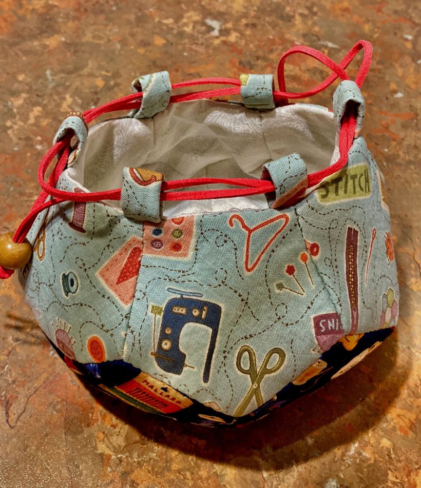 A small fabric bag in pale blue fabric with sewing motifs of scissors pins, stitched thread on it. The top closure is red cord as a draw string. The base show slightly in the corners where the pentagons meet, it has dark navy fabric with red and orange pattern. 