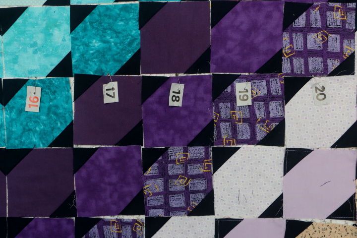 Snapshot of the design on the wall. Turquoise and purple half snowball blocks.  The numbers 16, 17, 18, 19, and 20 are pinned in order to different coloured blocks in preparation of sewing fabric blocks into columns .