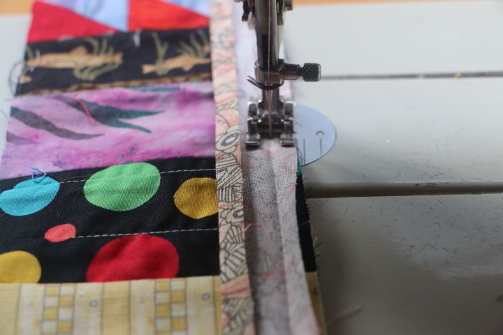 Close up of a sewing machine foot and needle, with fabric to the left side. The binding strip is being sewn to the fabric. The creased seam created by the tool and iron is a guide for the seam allowance.