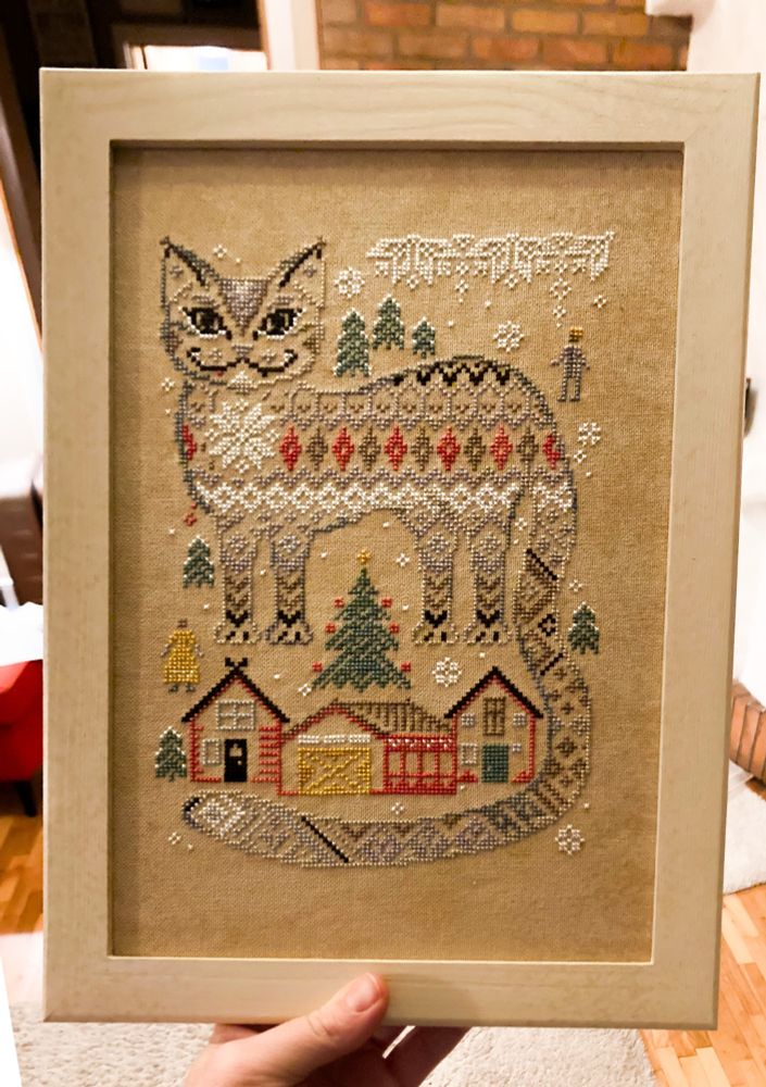 A vintage looking cross stitched image of a cat with Nordic motifs for fur, framed in a light wooden frame 