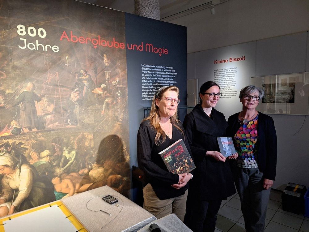 Ausstellungsteam (von links:) Friederike Koch-Heinrichs, Leiterin Museum der Westlausitz, Magdalena Schaffer, Archäologie-Volontärin und Autorin von "In Kamenz dunklen Gassen" und Susanne Hose, Kulturwissenschaftlerin und Volkskundlerin am Sorbischen Institut. Die Museumsleiterin hält den druckfrischen Ausstellungs-Katalog in den Händen. Im Hintergrund zu sehen ist das Titelmotiv der Ausstellung "800 Jahre Aberglaube und Magie": die "Hexenversammlung" von Frans II. Francken (1581-1642, Antwerpen), datiert 1607, ausgestellt im Kunsthistorischen Museum in Wien © Sorbisches Institut (2025)