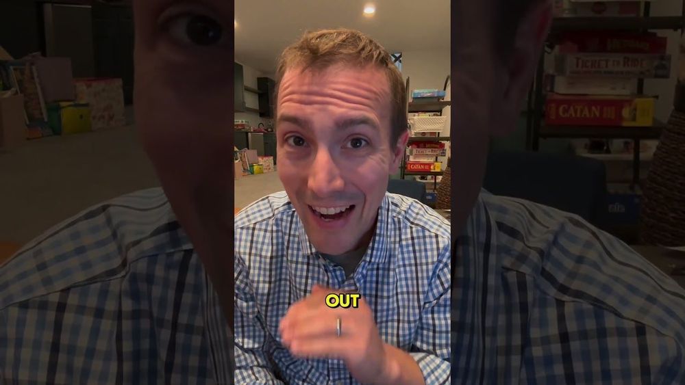 A white man with short hair and a checked shirt sits in what looks like a large finished basement with boxes on shelves in the background, speaking earnestly to his phone camera.