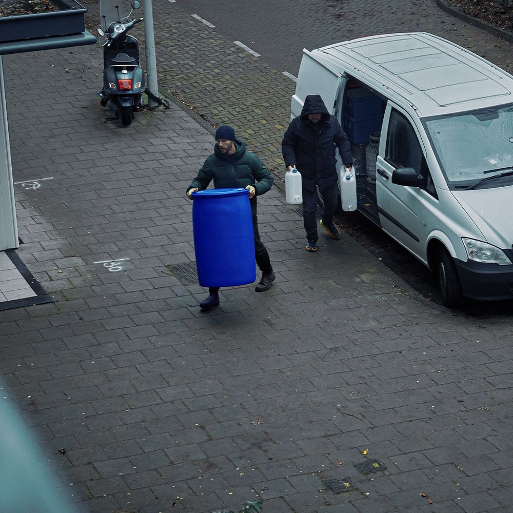 Beeld uit de campagne 'Vreemd of verdacht. Houd misdaad uit je buurt.' Twee mannen dragen een blauwe ton en twee witte jerrycans vanuit een bestelbusje naar een gebouw.