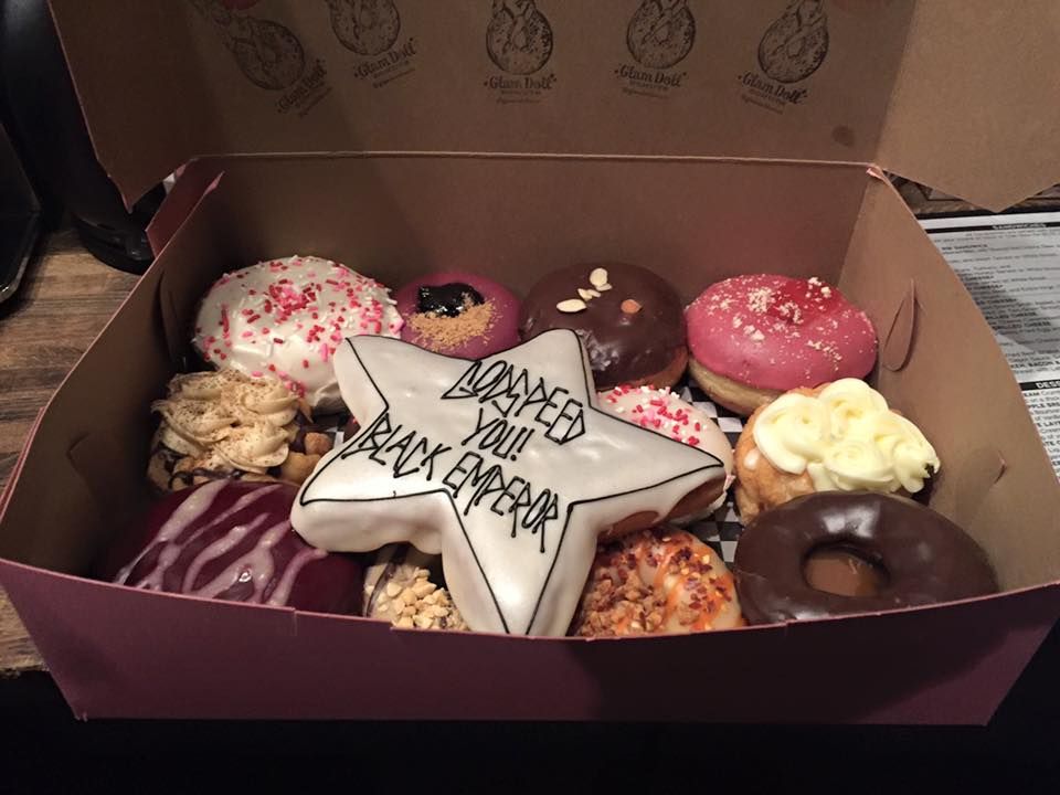 A box of assorted donuts, the one on top has "Godspeed You! Black Emperor" written in icing. The donut with the writing is star shaped and the writing is black on silver/white icing