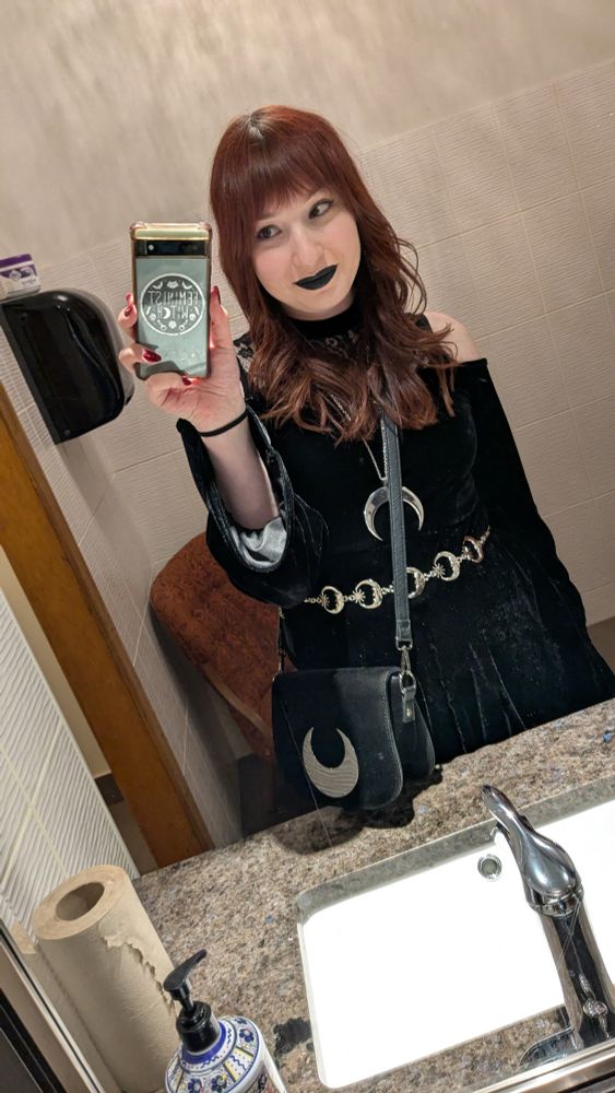 It's Danielle! She's wearing a black velvet dress with cold shoulders and long sleeves,  black lipstick, and several pieces of silver moon jewelry and accessories. she's in a washroom in this picture. 