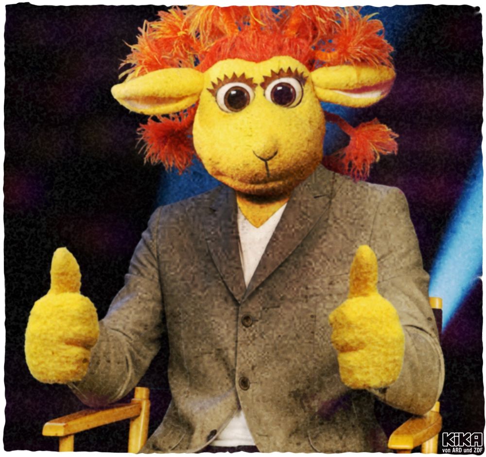 Chili das Schaf sitzt in einem Regiestuhl und zeigt beide Daumen nach oben 