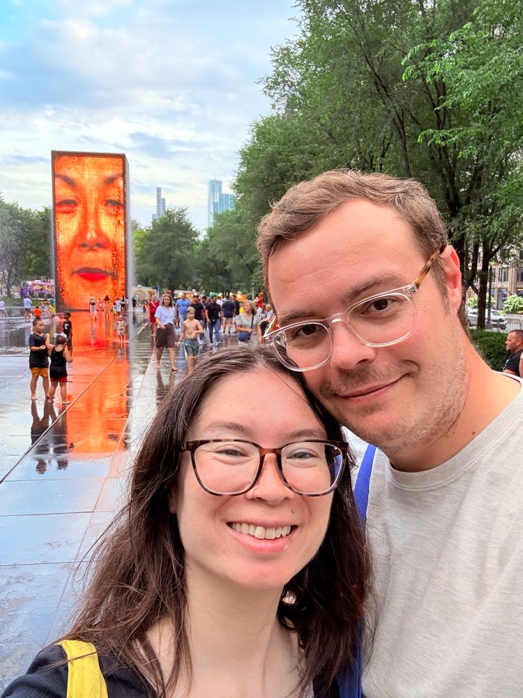 A smiling couple taking a selfie in front of Crown Fountain in Chicago’s Millennium Park. Behind them, one of the fountain’s large LED towers displays a close-up face of a woman, with water visibly running down. The park is busy with people and surrounded by trees and tall buildings in the background.