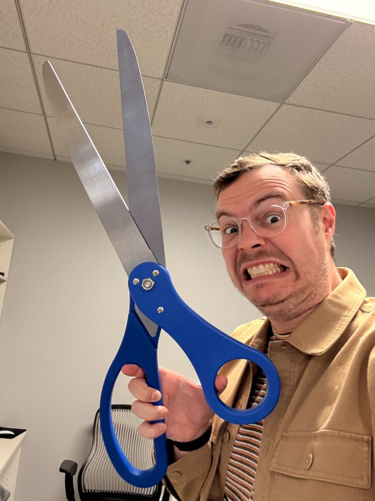 A man wearing glasses and a tan jacket makes a comically exaggerated grimace while holding an oversized pair of novelty scissors with blue handles and large silver blades. He is indoors in an office setting with a ceiling of acoustic tiles and fluorescent lighting, and a mesh-back office chair is visible in the background.