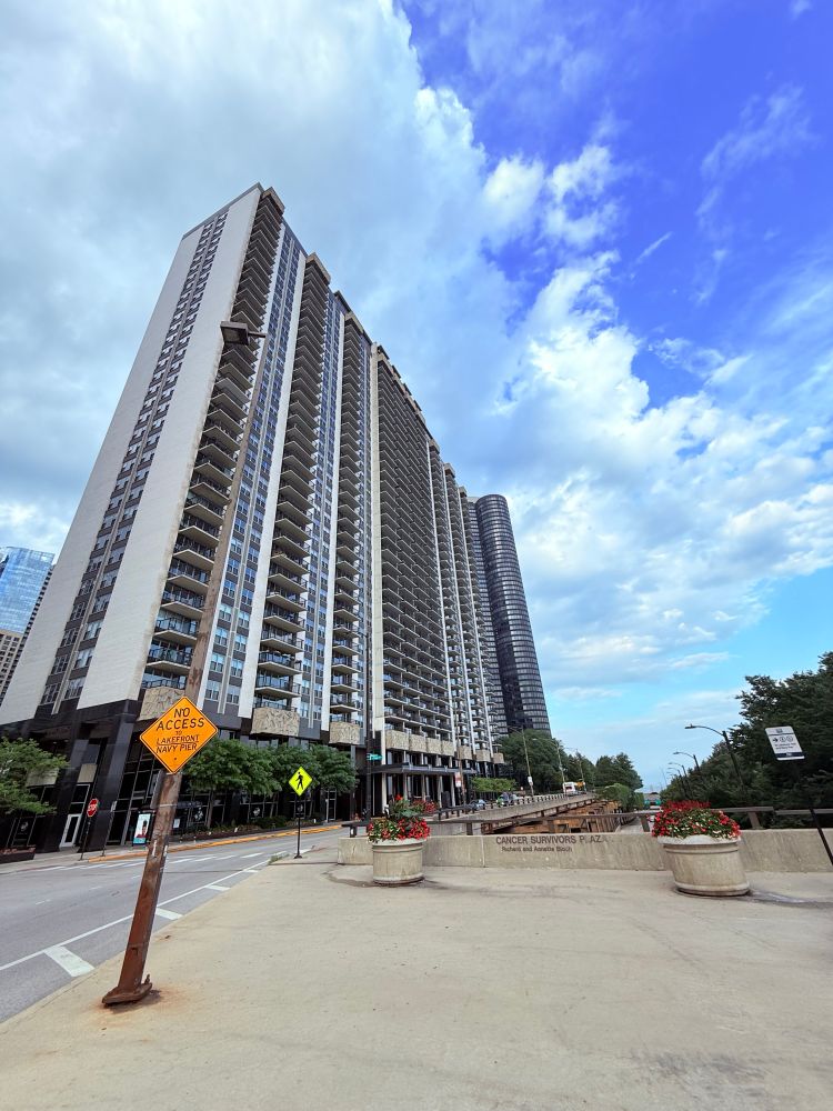 Tall residential high-rise buildings with numerous balconies viewed from a low angle, under a partly cloudy blue sky. A signpost in the foreground reads “NO ACCESS LAKEFRONT NAVY PIER,” and large concrete planters with red flowers line a pathway marked “CANCER SURVIVORS PLAZA.”
