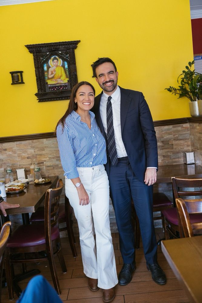 Alexandria Occasio-Cortez and Zohran Mamdani stand side by side smiling in a brightly lit restaurant with a yellow wall. The woman wears a blue and white striped blouse tucked into high-waisted white pants and brown boots. The man is dressed in a dark suit with a white shirt and striped tie. Behind them, a framed image of Buddha hangs on the wall, along with a potted plant and several wooden chairs and tables set with plates and drinks.