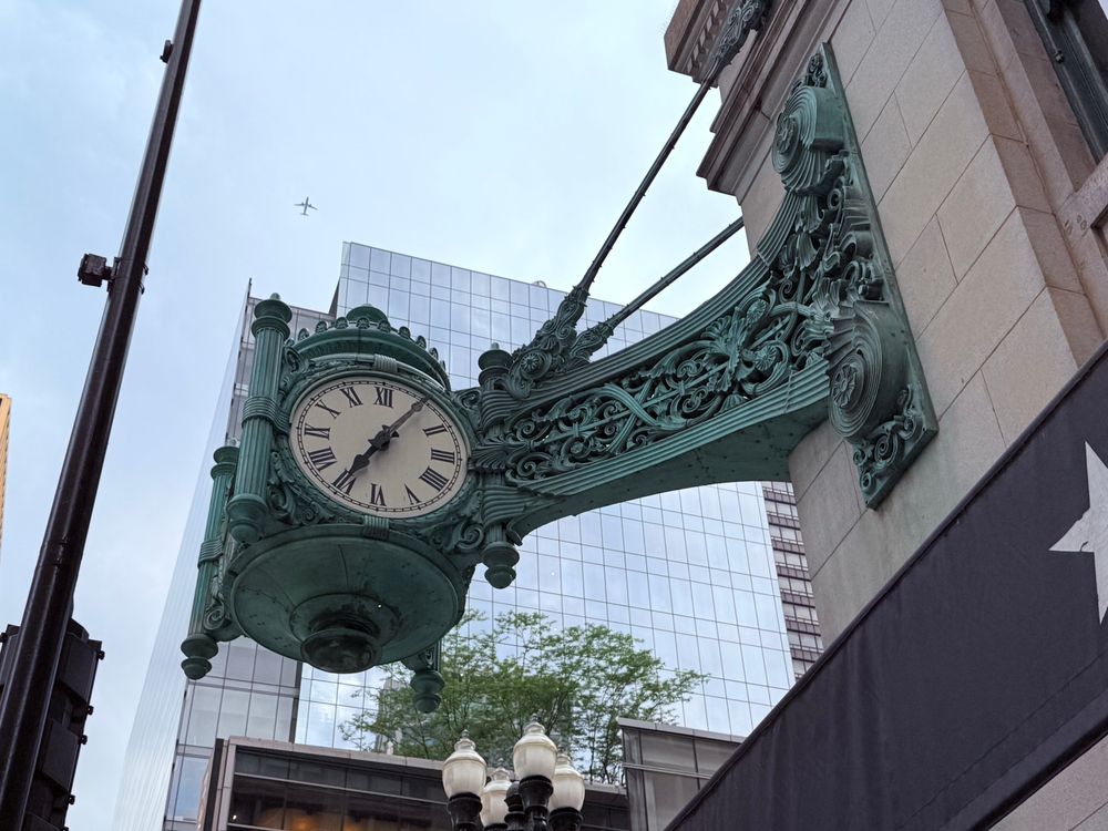 A close-up view of the historic green ornamental clock mounted on the corner of a building, displaying Roman numerals with the time at approximately 5:39. Behind the clock, modern glass skyscrapers and a faint airplane in the sky are visible.