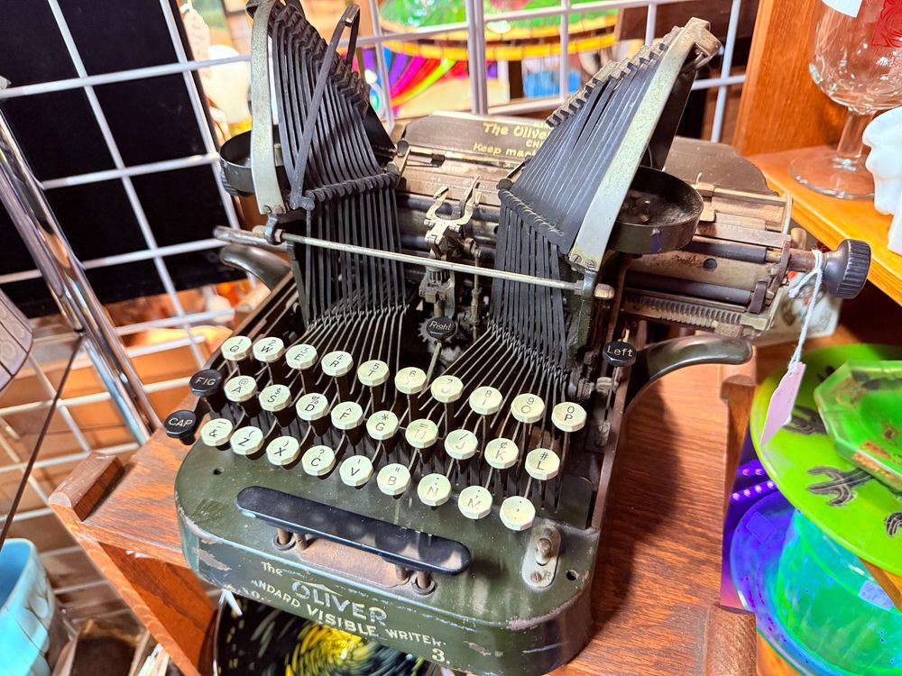 Close-up of a vintage Oliver typewriter with an olive green metal body and raised typebars arranged in an inverted “U” shape on both sides. The keys are round with white tops and black letters, including labels like “FIG” and “CAP” on the sides. The front reads “The OLIVER STANDARD VISIBLE WRITER.” The typewriter sits on a wooden shelf among colorful glassware and other vintage items.