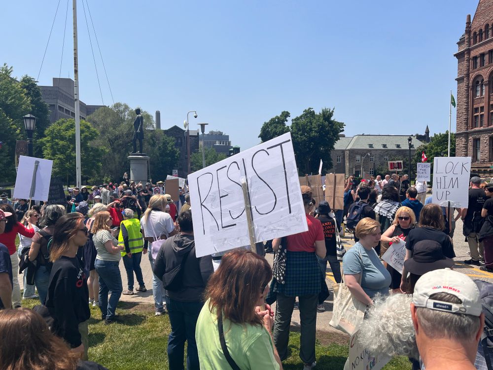 A crowd protesting, many carrying American/Canadian flags or signs. A RESIST sign is prominent in frame. 