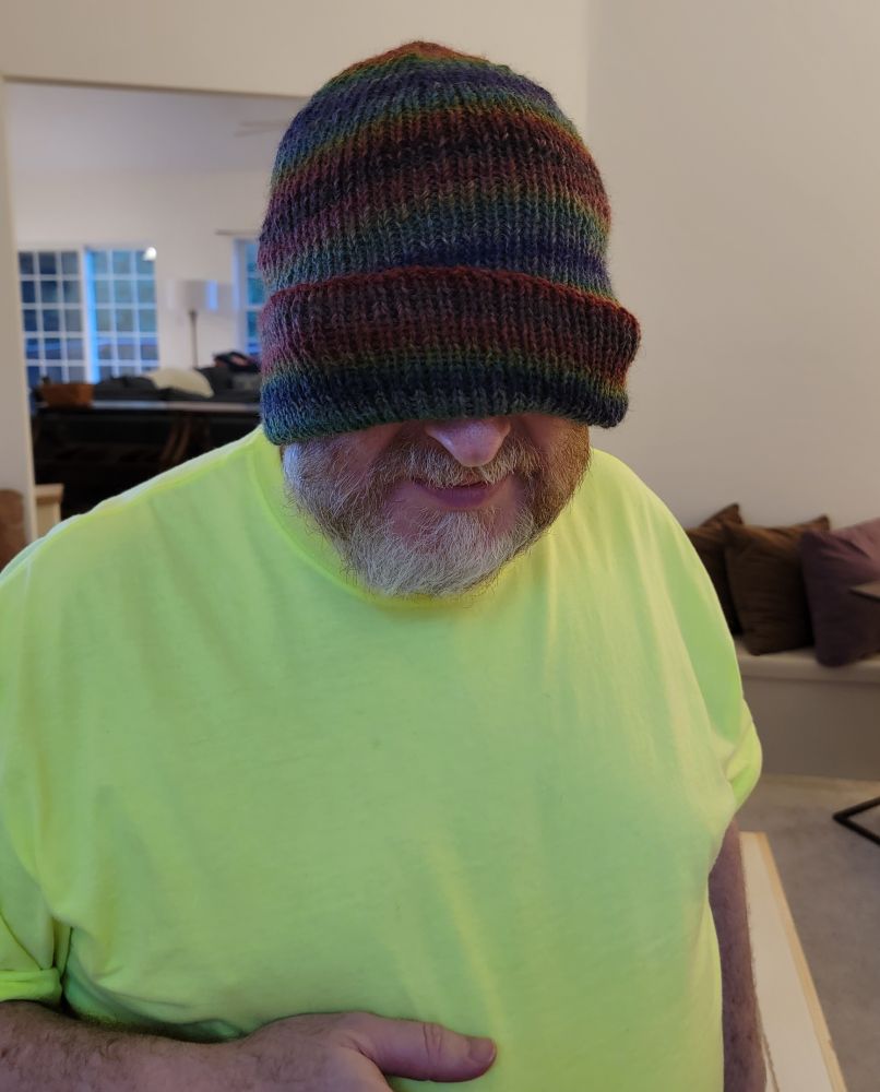 A bearded man wearing a safety yellow t-shirt and a striped handknit hat which is pulled down over his eyes.