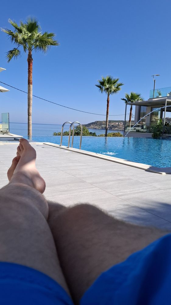 Männerbeine in Badehose vor einem Infinity-Pool mit Palmen. Im Hintergrund ist das Meer zu sehen.