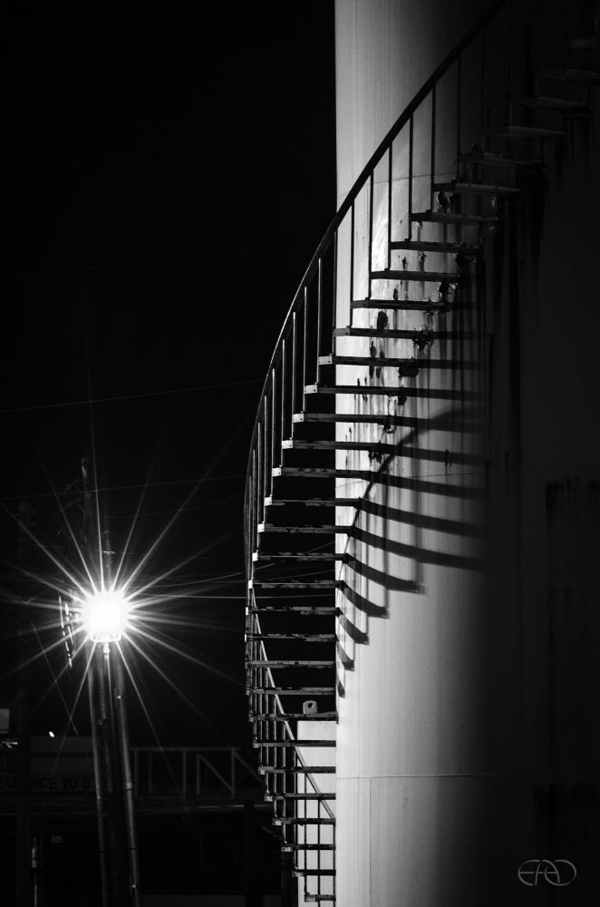 A monochrome night shot of an industrial cylindrical tank with a curved stair wrapping around it. The lamp post beyond casts shadows across the curved surface of the tank.