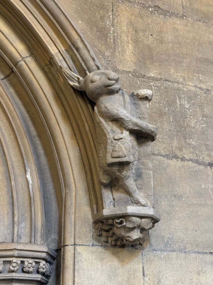 Mediaeval carving of a hare standing upright with a satchel, at the end of a stone arch moulding, with the archway to the left.