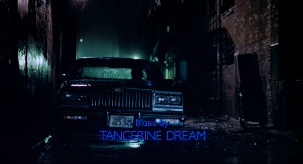 screenshot from Thief featuring a black Buick Regal parked in a narrow alley at night, backlit by a green glowing light. at the bottom of the screenshot, it reads 'Music by TANGERINE DREAM' in blue letters