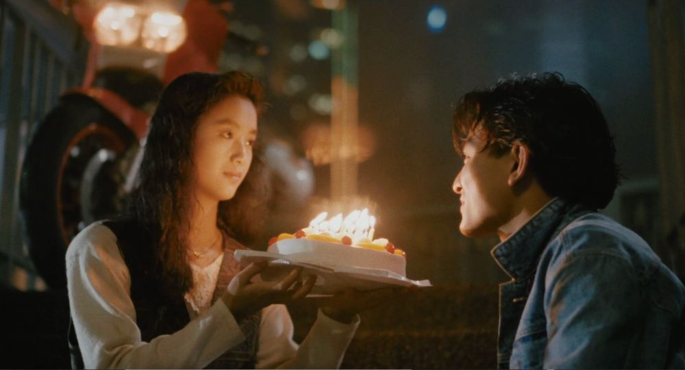 Chien Lien Wu (wearing dark vest over long-sleeve white dress) and Andy Lau (with acid-wash denim jacket + popped collar) sitting outside about to share a birthday cake that she's holding. his Suzuki is out of focus in the background to the left. 