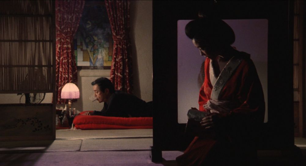 wide shot of Tatsuya Nakadai smoking on the floor near a little lamp on the left with a geisha sitting in front of a screen on the right 