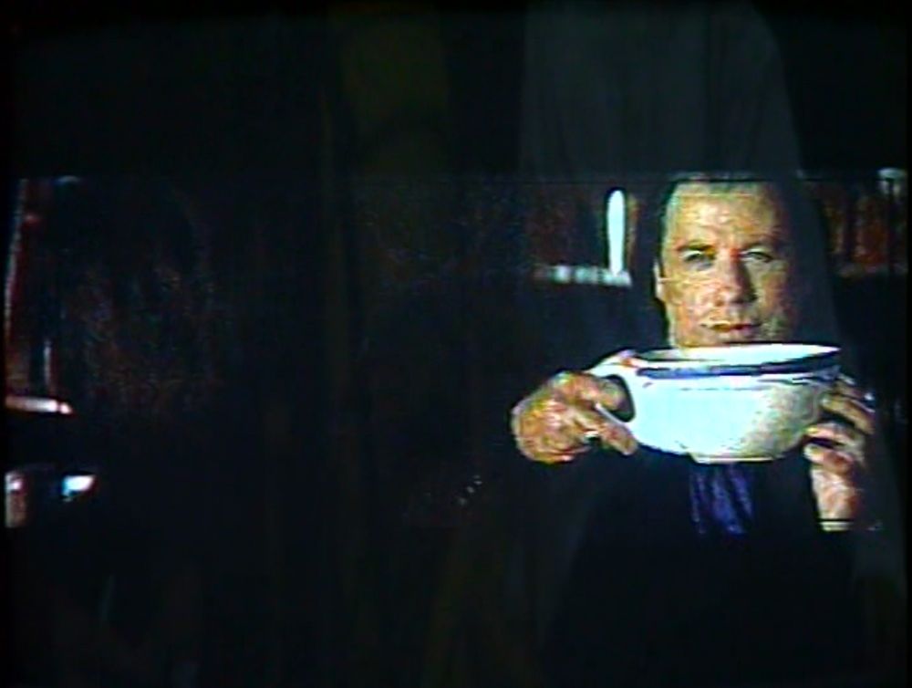 blurry/low res shot of Travolta keeping it together but looking a little smug as he holds an enormous white teacup that appears to be slightly bigger than his head. 