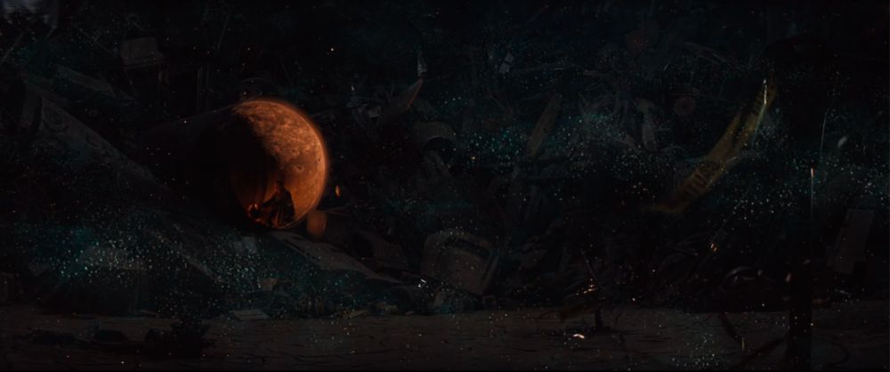 a dissolve: a wide shot of Kurt Russell's Todd sitting inside a rusty open pipe/tunnel, surrounded by desert and piles of garbage, with the overlapping image of the night sky: a rust-colored planet and a bluish blanket of stars 