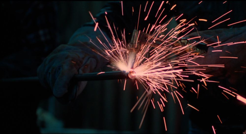closeup on a gloved hand holding a metal rod, the other hand holding a blowtorch being applied to the metal creating a spray of pinky gold sparks 