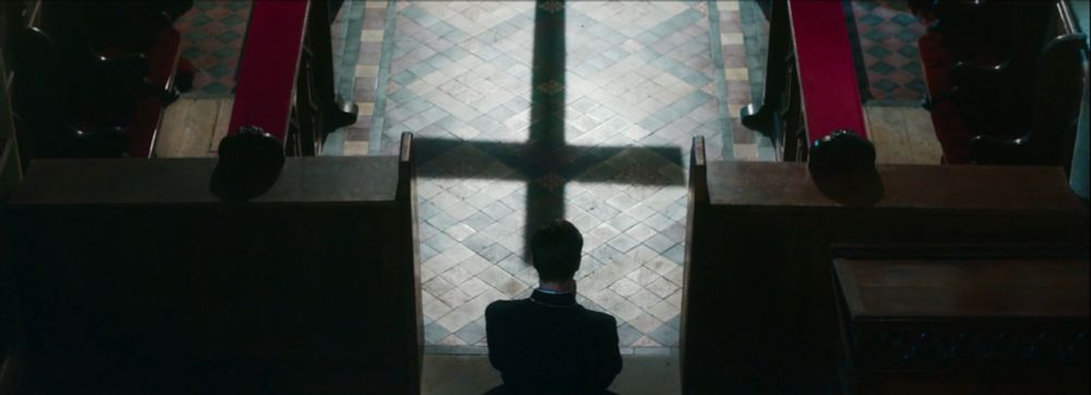 overhead shot of minister - seen from behind - kneeling and praying between pews in church. a cross casts a long upside-down shadow on the tile floor in front of him. 