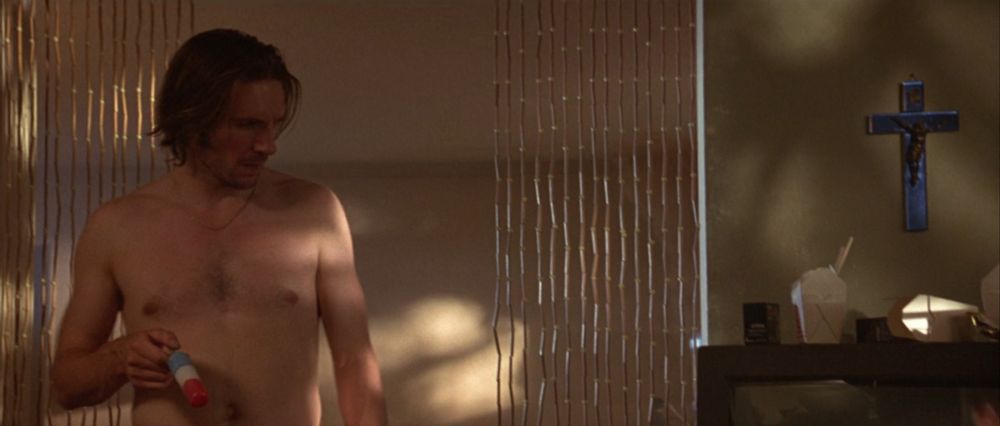 incredibly beautiful shot of shirtless Fiennes walking into his living room, illuminated by mottled sunlight coming in through the window, absently dangling his firecracker popsicle (red, white, & blue) as he looks down at the TV. there's a beaded curtain behind him and a silvery blue crucifix on the wall behind the TV. 