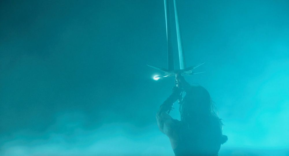 wide shot of Bernhardt seen from behind wielding giant 4-bladed sword, swathed in aqua blue smoke/fog  