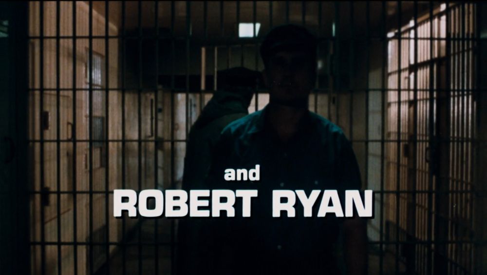title card for The Outfit that reads 'and ROBERT RYAN' in large white font over a shadowy Robert Duvall walking through prison 